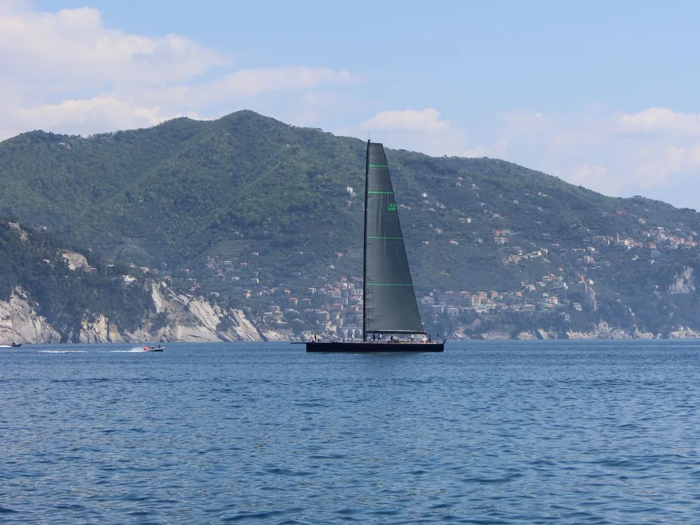 Dark-hulled sailing yacht under sail beside a mountainous coast